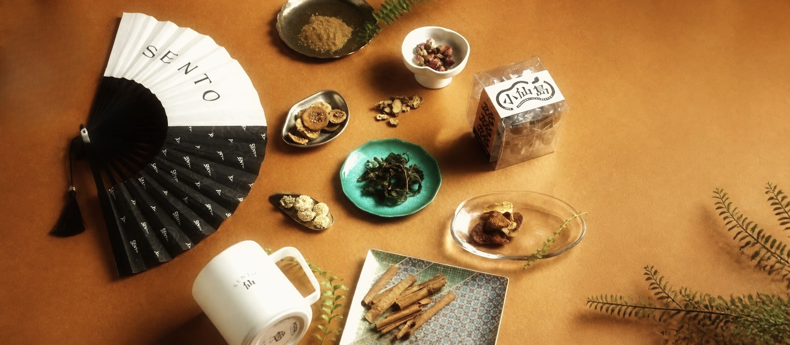 小仙島漢方手搖飲的原料：玫瑰花、陳皮、肉桂、茶葉與小仙島茶包，旁邊是 SENTO 摺扇、馬克杯與蕨葉